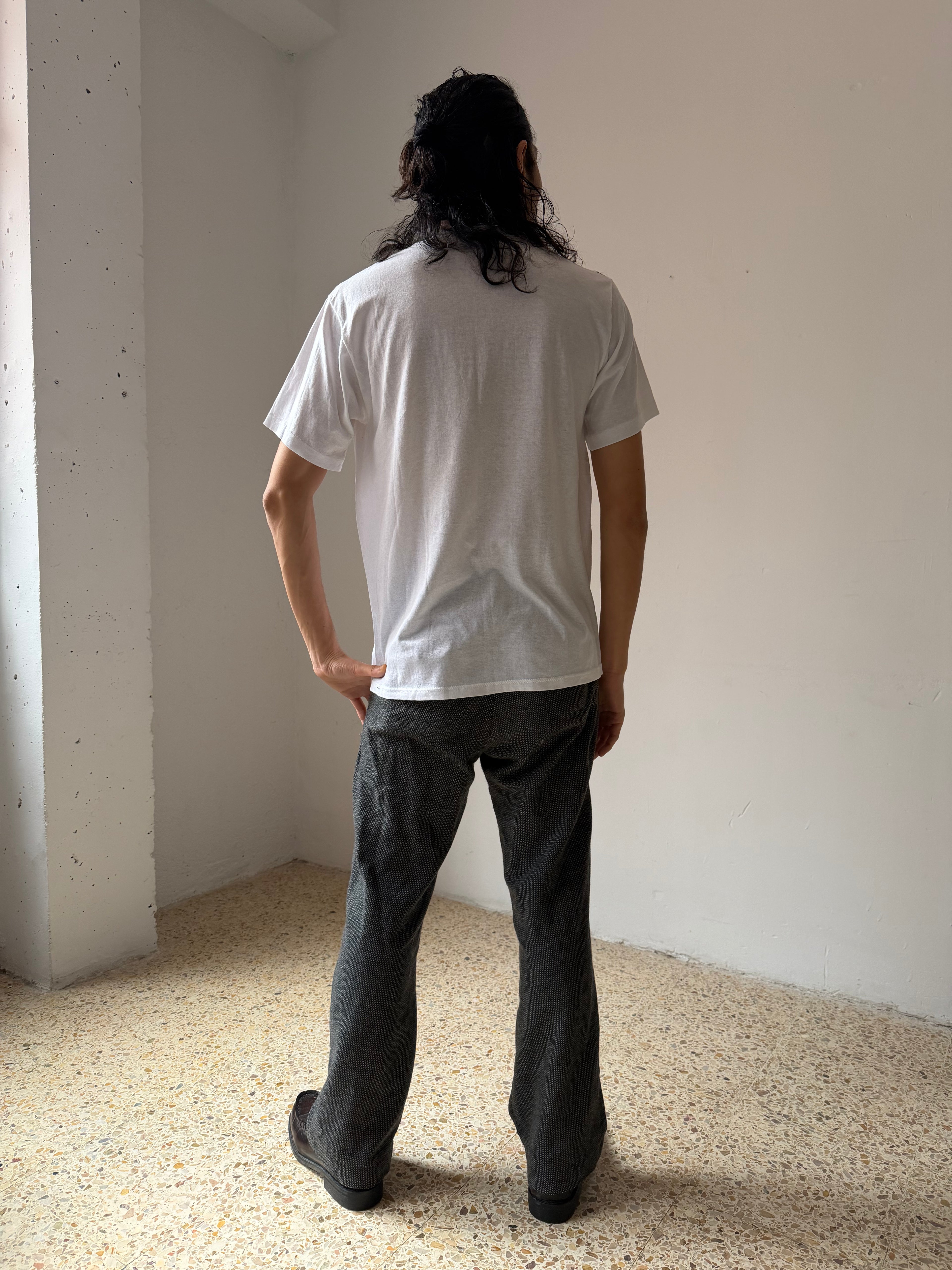 Person wearing a white t-shirt and dark pants standing in a minimalistic room with light-colored floor and walls.