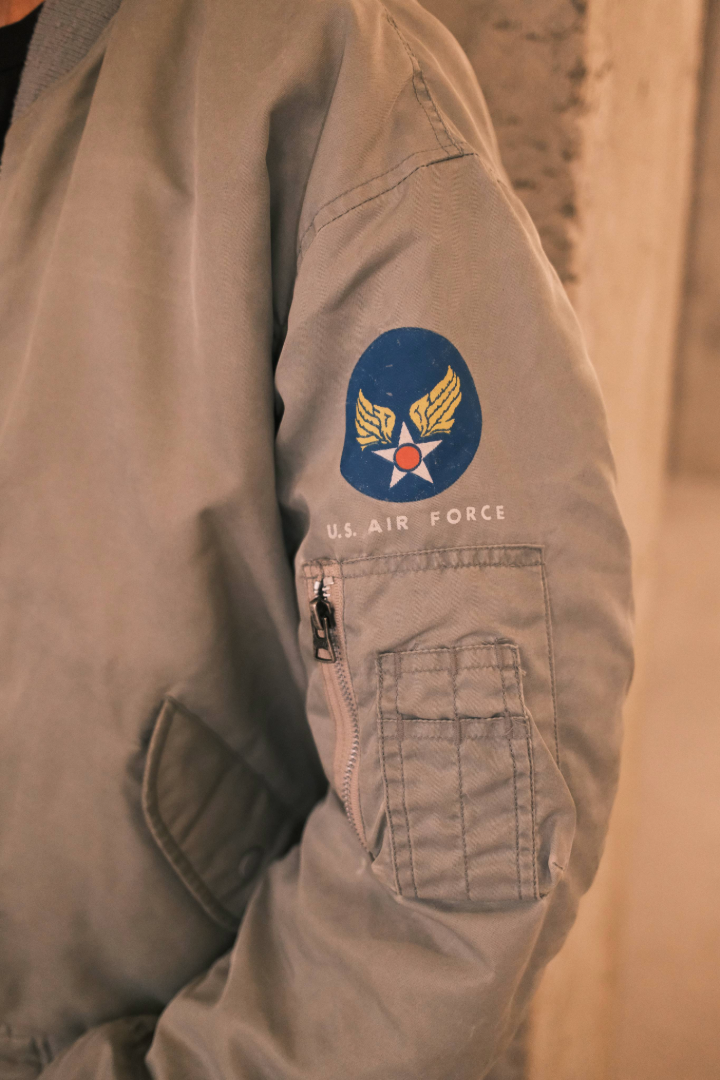 Khaki flight jacket with a patch on the sleeve against a neutral background