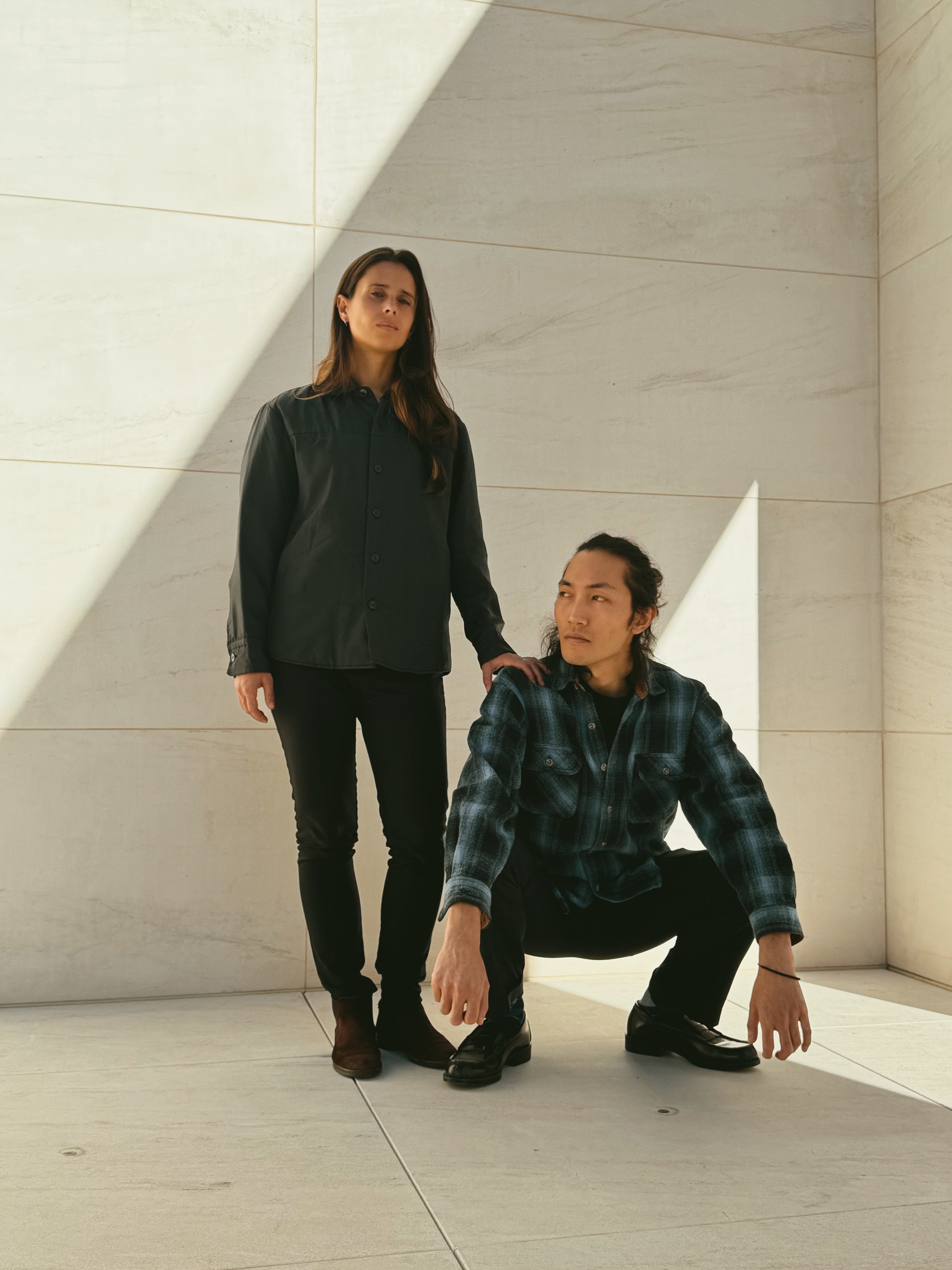 Two people in dark clothing standing against a light-colored wall.