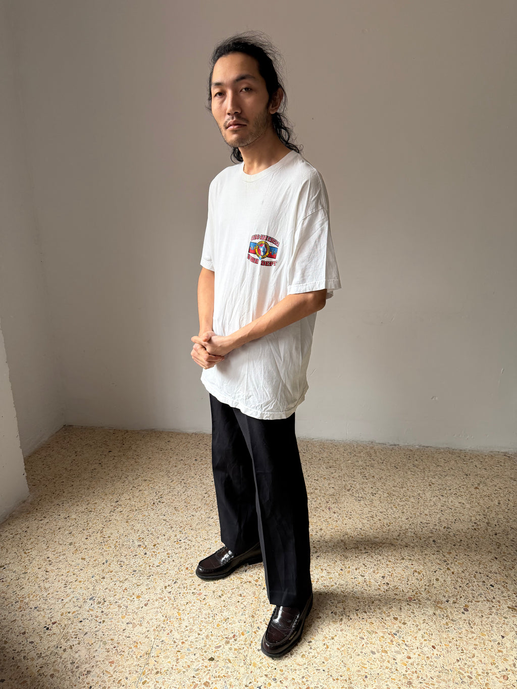 Man wearing a white t-shirt with a colorful graphic and black pants standing in a minimalistic room.