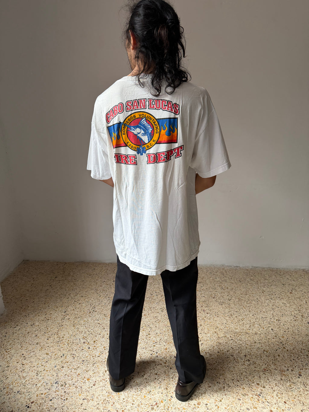 Person wearing a white t-shirt with colorful graphics and text, standing in a room with a plain background.