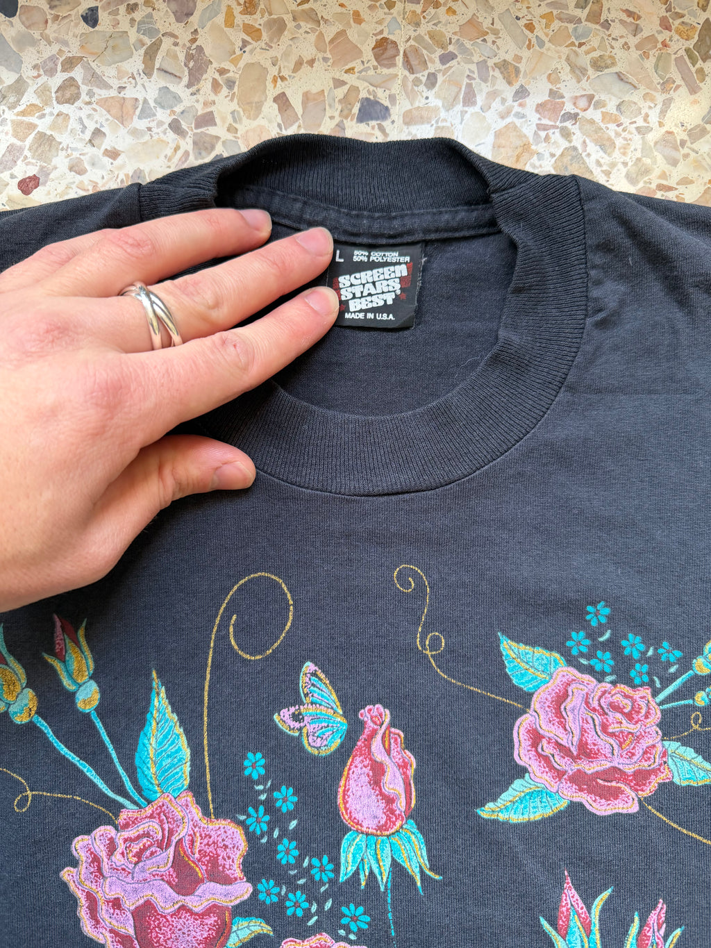 Dark blue t-shirt with floral and butterfly designs held by a hand on a terrazzo background