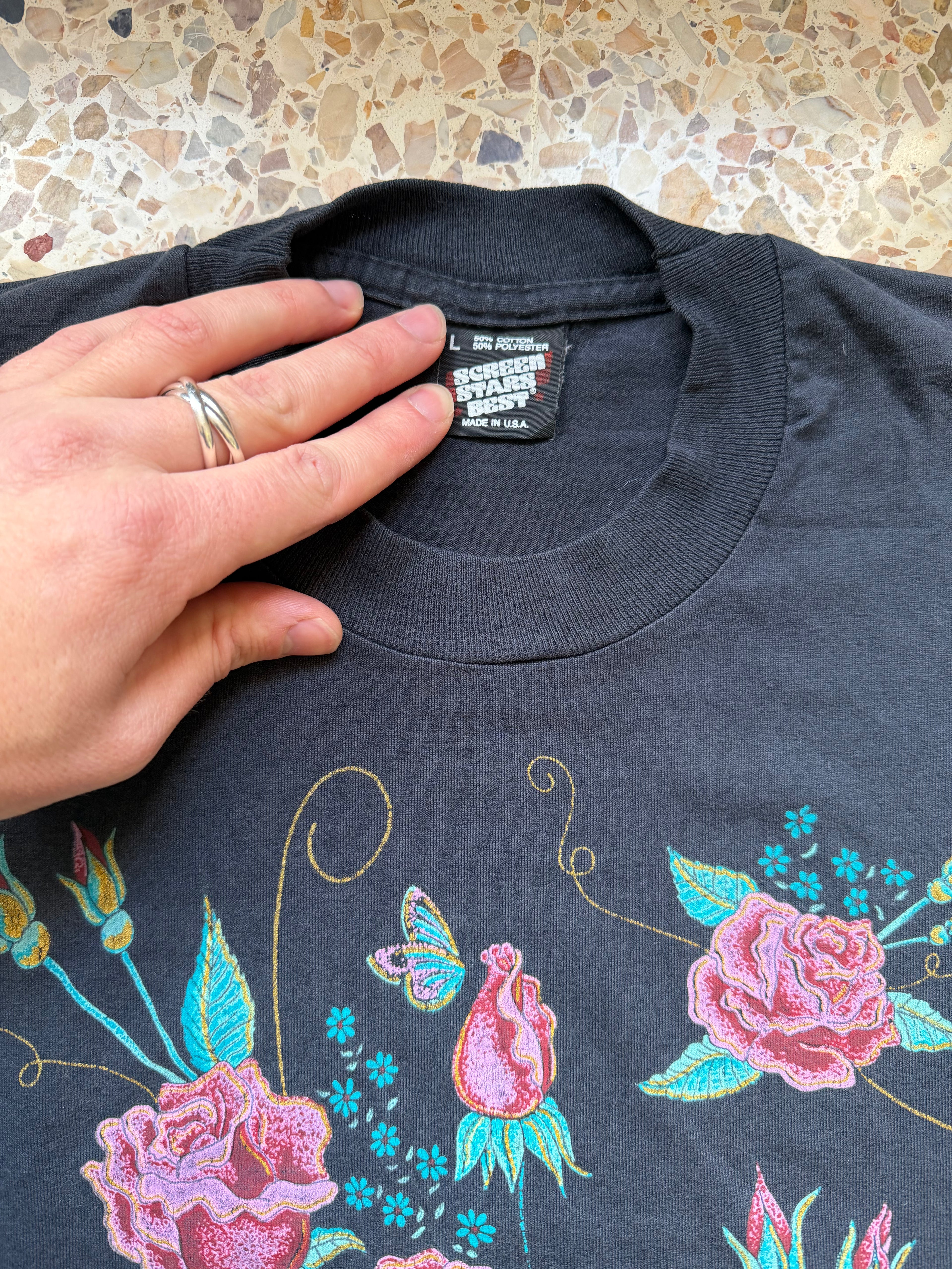Dark blue t-shirt with floral and butterfly designs held by a hand on a terrazzo background