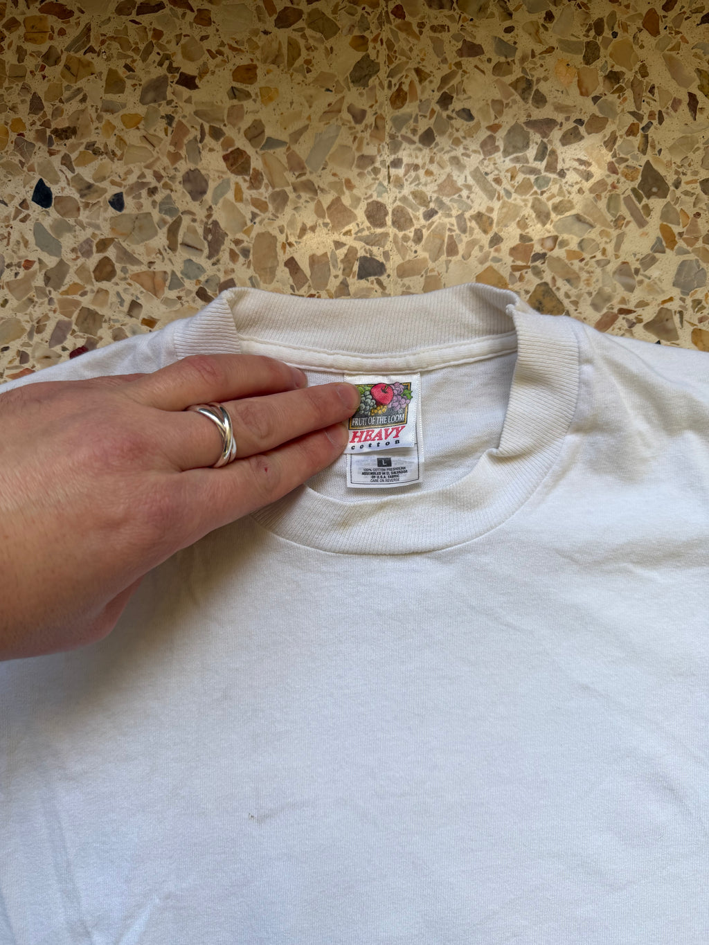 White t-shirt with a label held by a hand against a terrazzo wall.