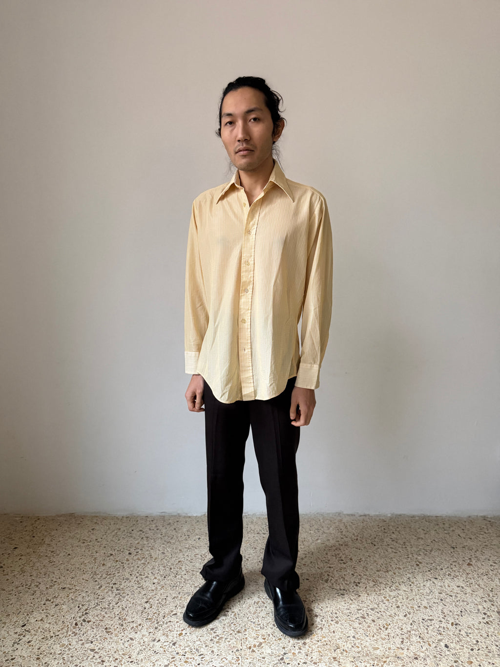 Person wearing a light-colored shirt and dark pants standing against a plain background