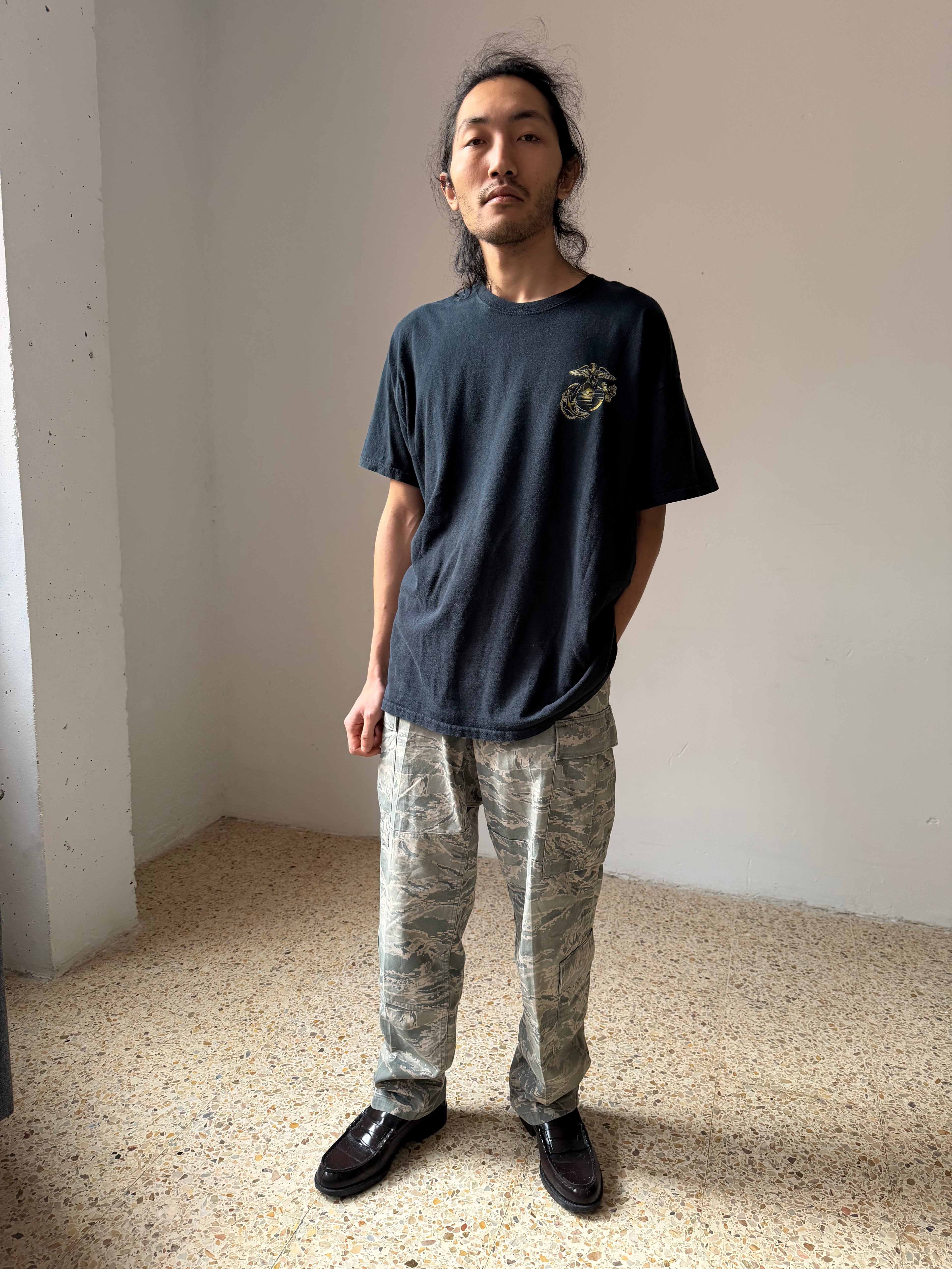 Person wearing a black Marines t-shirt and camouflage pants standing in a minimalistic room.