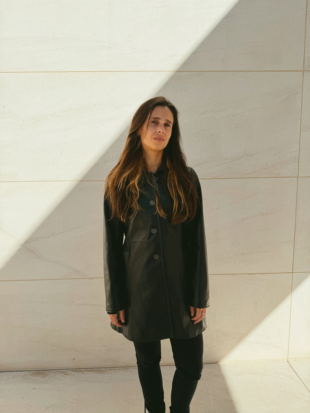 Woman wearing a Balmain black leather coat standing against a light-colored wall.