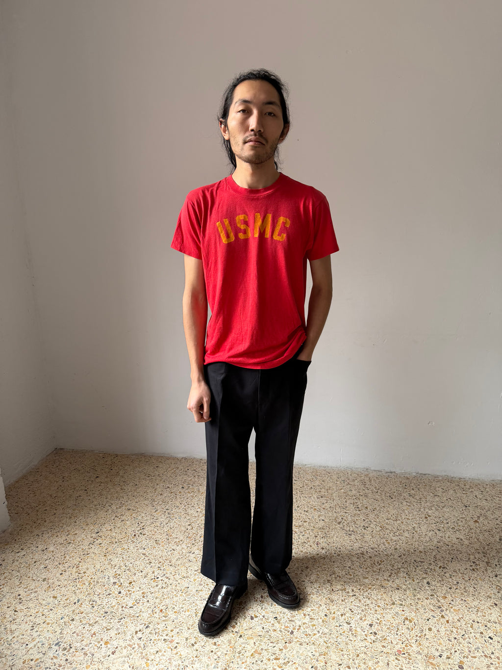 Person wearing a red t-shirt with yellow usmc text standing in a minimalistic room.