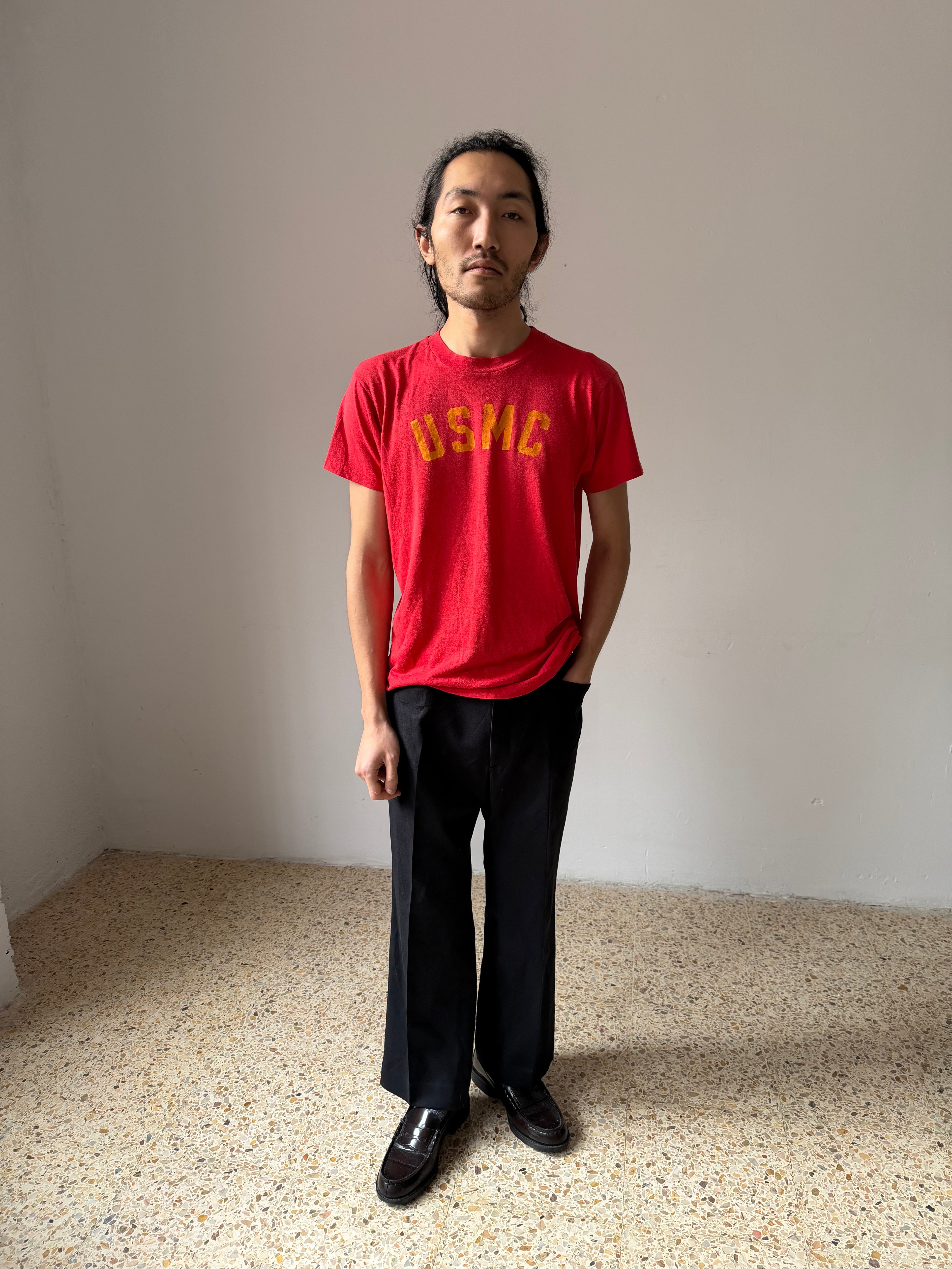 Person wearing a red t-shirt with yellow usmc text standing in a minimalistic room.